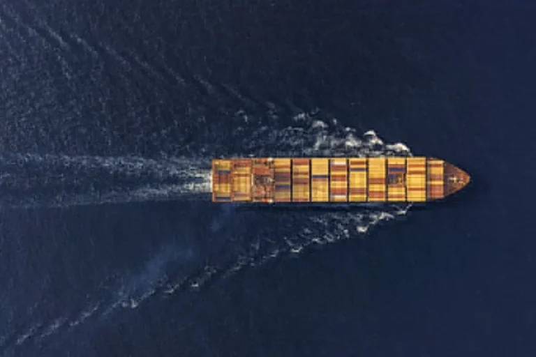 Aerial view of a cargo container ship sailing in dark blue water, leaving a wide wake behind it.