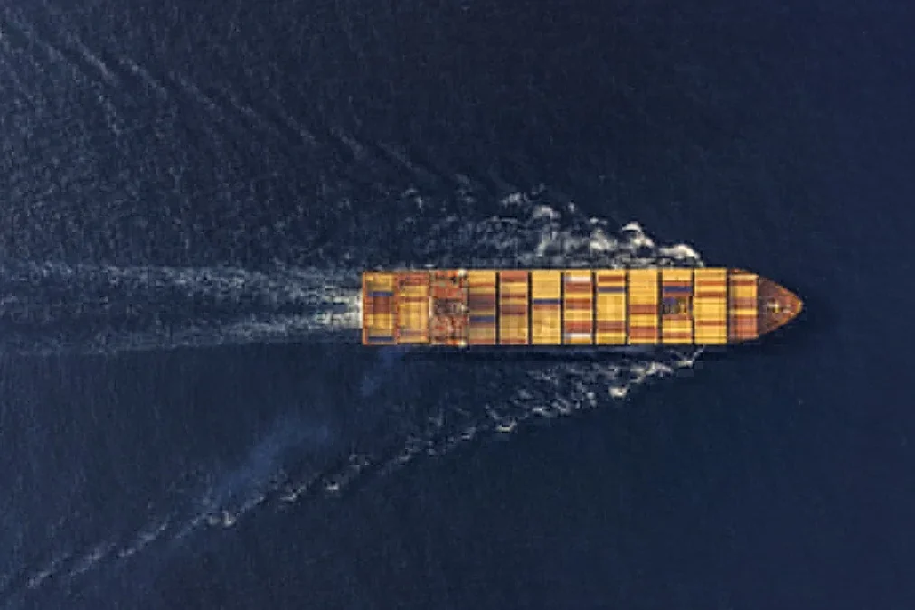 Aerial view of a cargo container ship sailing in dark blue water, leaving a wide wake behind it.