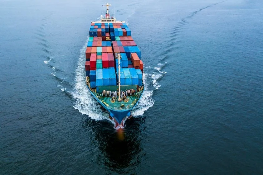 Top-down view of a cargo ship at sea, with colorful shipping containers stacked on deck and a visible wake behind it.